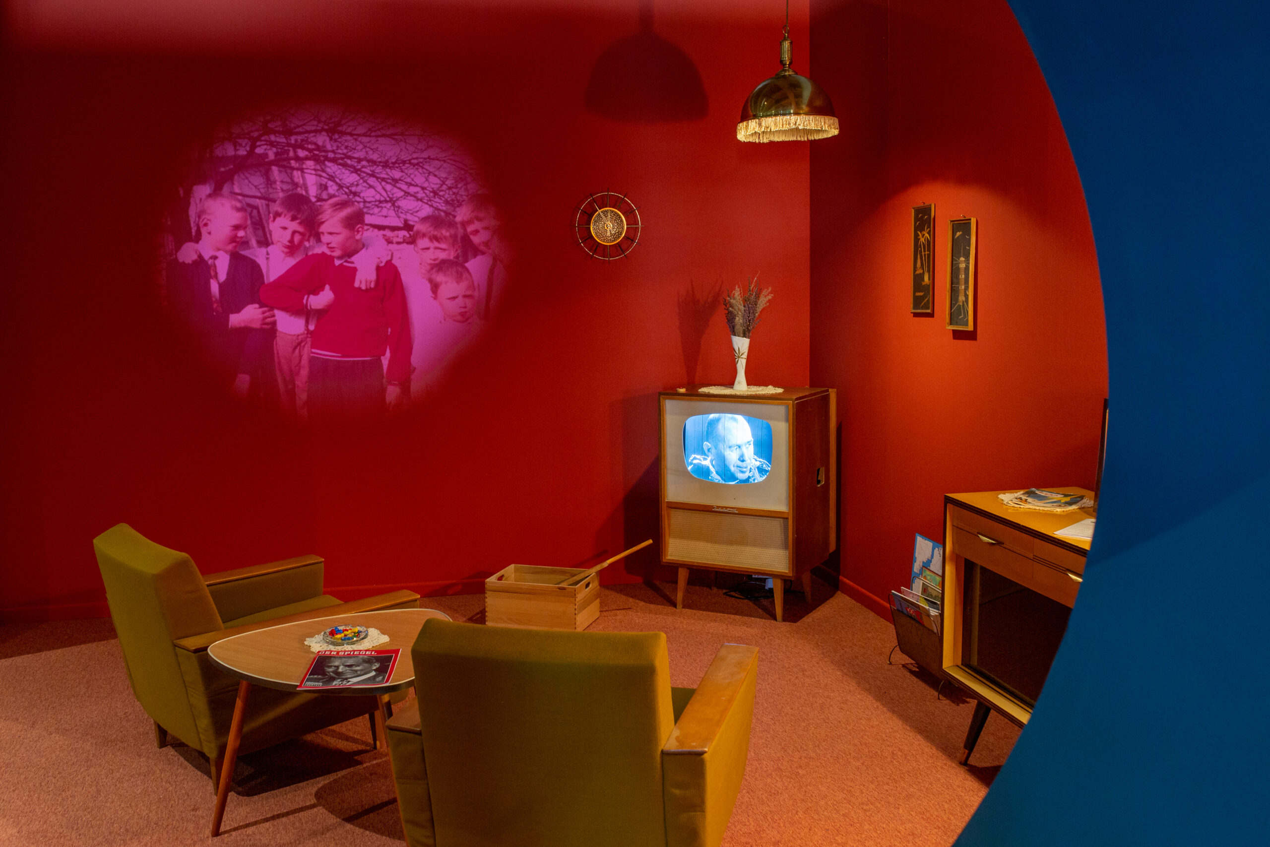 Nachgestelltes 1960er-Jahre-Wohnzimmer mit roten Wänden, zwei grünen Retro-Sesseln, einem runden Holztisch, altem Fernseher mit Schwarz-Weiß-Bild, Projektionsfoto von Kindern an der Wand sowie Sideboard und Deckenlampe.