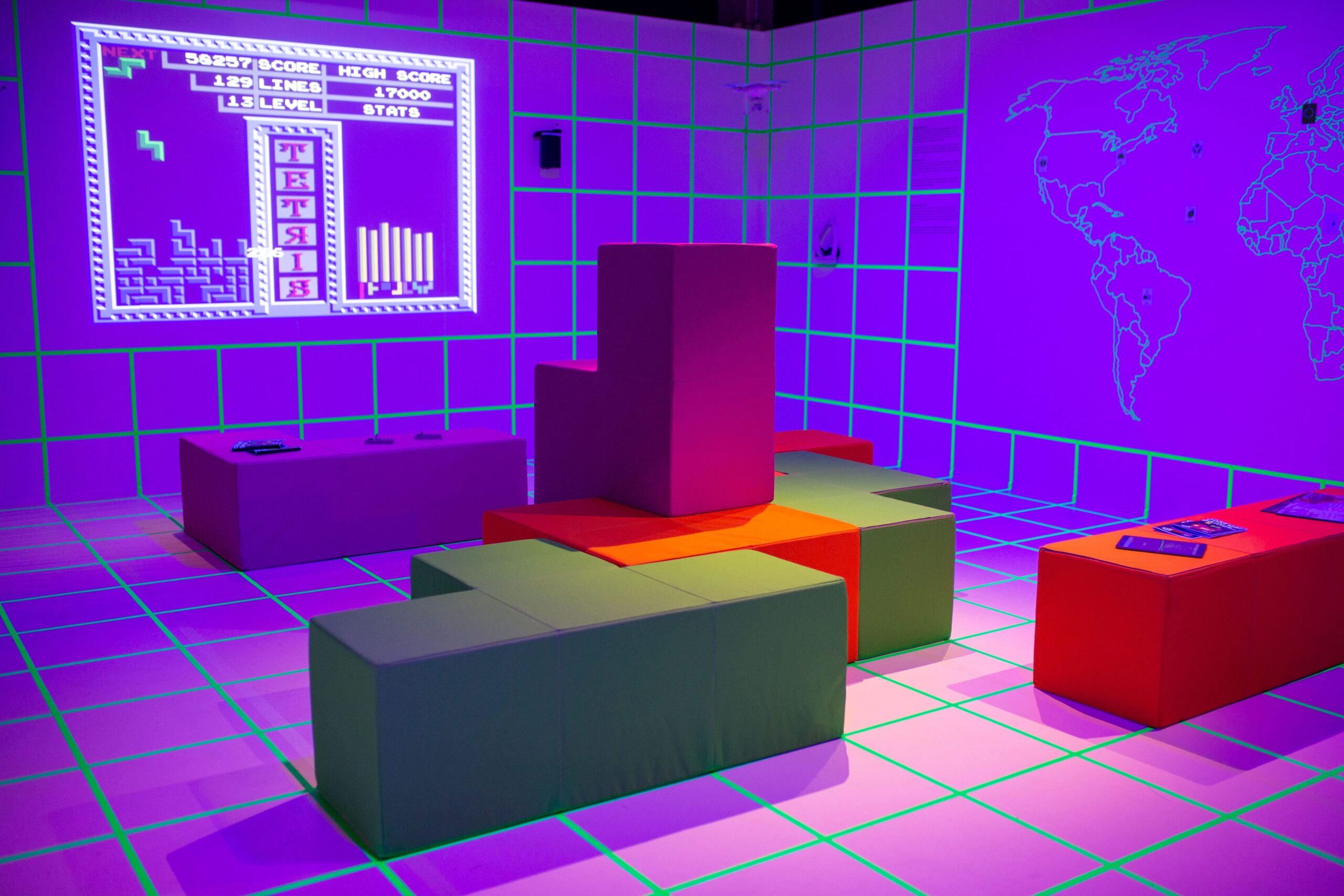 Beleuchteter Raum mit neonfarbenem Raster, Tetris-Projektion an der Wand und farbigen Sitzwürfeln in Form von Tetris-Steinen; daneben eine Weltkarte und ein Tisch mit Informationsmaterial.