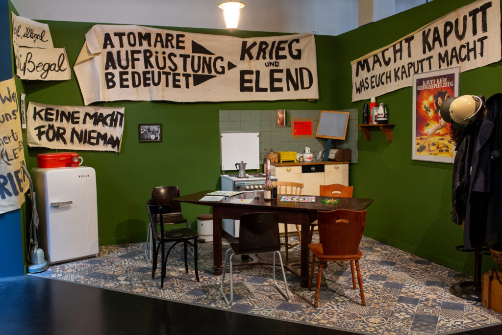 <p>Cuisine d’activistes reconstituée des années 1970/80, avec des banderoles de protestation, des meubles rétro, une table de cuisine et divers objets du quotidien</p>