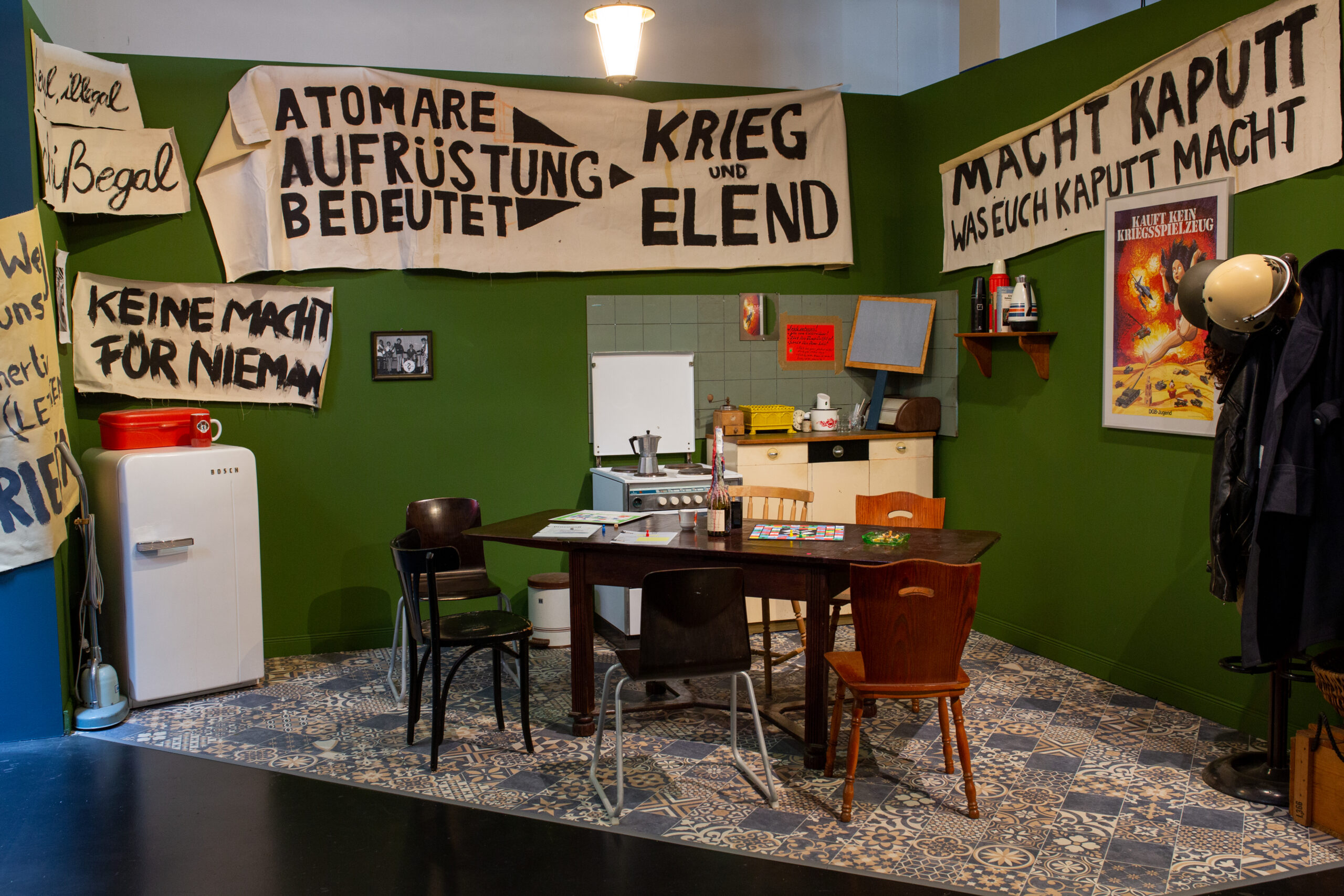 Das Bild zeigt eine nachgestellte, wohnlich wirkende Szene, die stark an eine politische WG oder Aktivisten-Küche aus den 1970er/80er Jahren erinnert