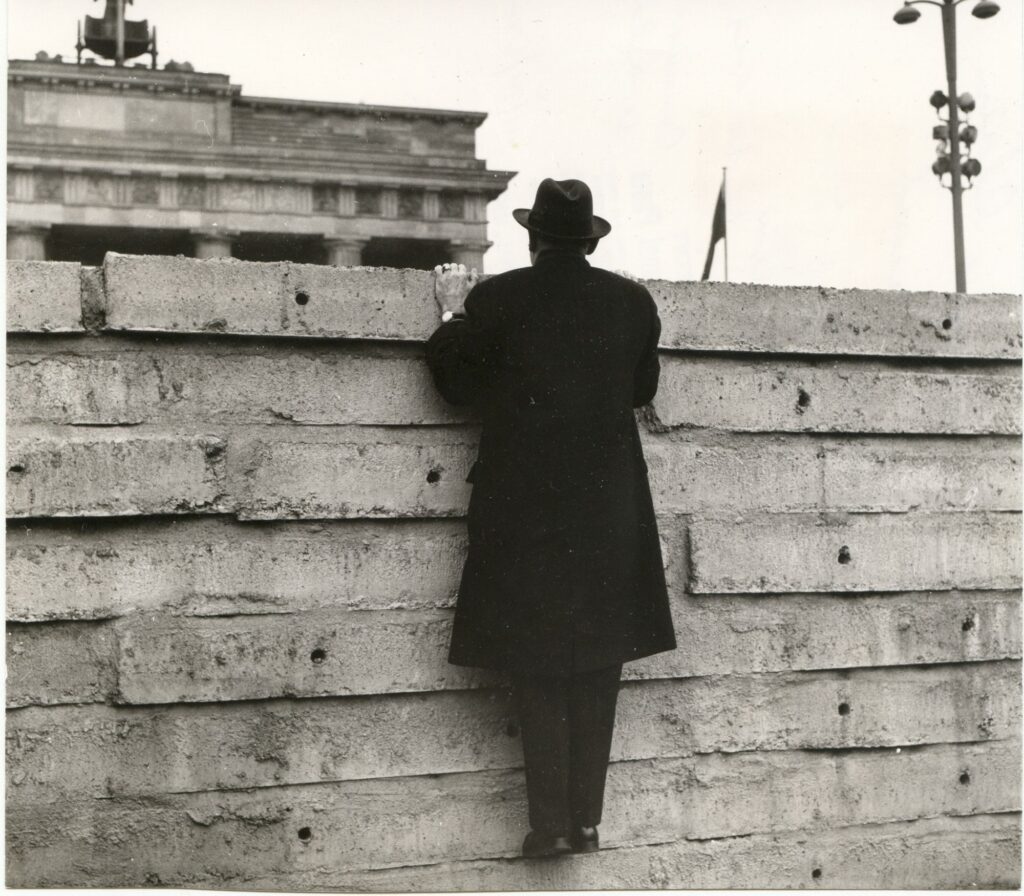 Lucius D. Clay zieht sich an der Berliner Mauer hoch und blickt auf das Brandenburger Tor.