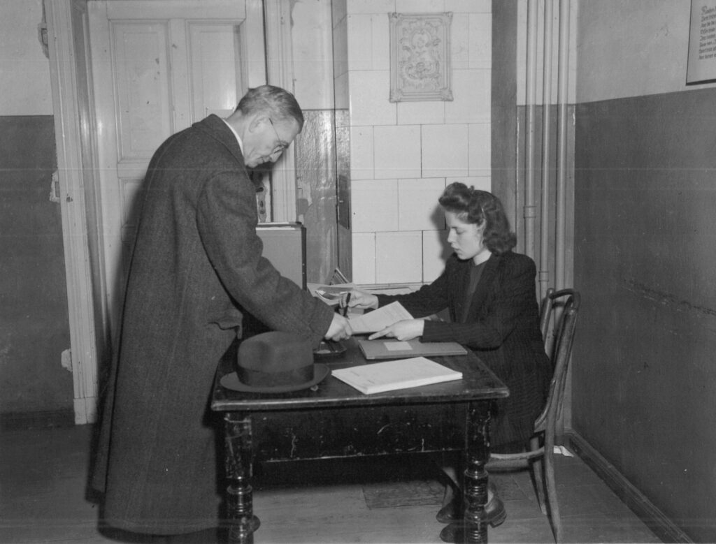 A man stands in front of a table and hands a denazification questionnaire to the woman sitting behind it.