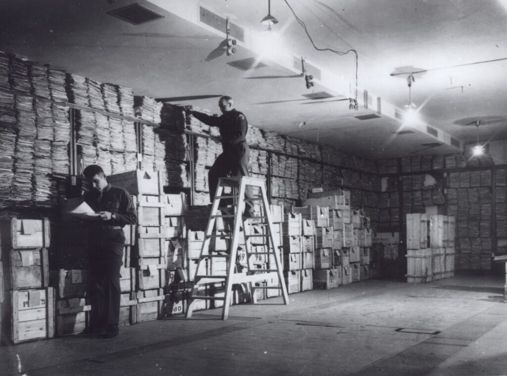 Two soldiers in a windowless room with documents and boxes piled high.