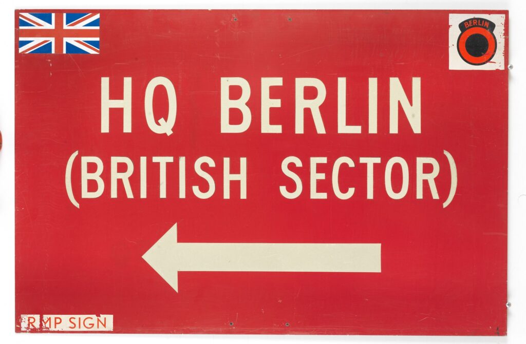 Panneau directionnel rectangulaire rouge, avec flèche et inscription blanches « HQ Berlin (British Sector) » indiquant le quartier général berlinois dans le secteur britannique