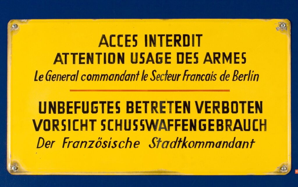 Rectangular yellow warning sign with an inscription in black in German and French reading “No Access for Unauthorized Persons. Caution Firearms in use. The Commandant of the French Sector. / Accès interdit. Attention usage des armes. Le Général commandant le Secteur Français de Berlin“