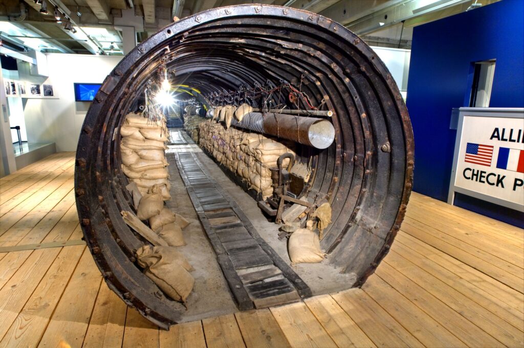 Vue de l’intérieur du tunnel d’espionnage composé d’un tuyau en métal avec, sur les deux côtés, des sacs de sable, des câbles et, au centre, une rangée d’ampoules et un passage