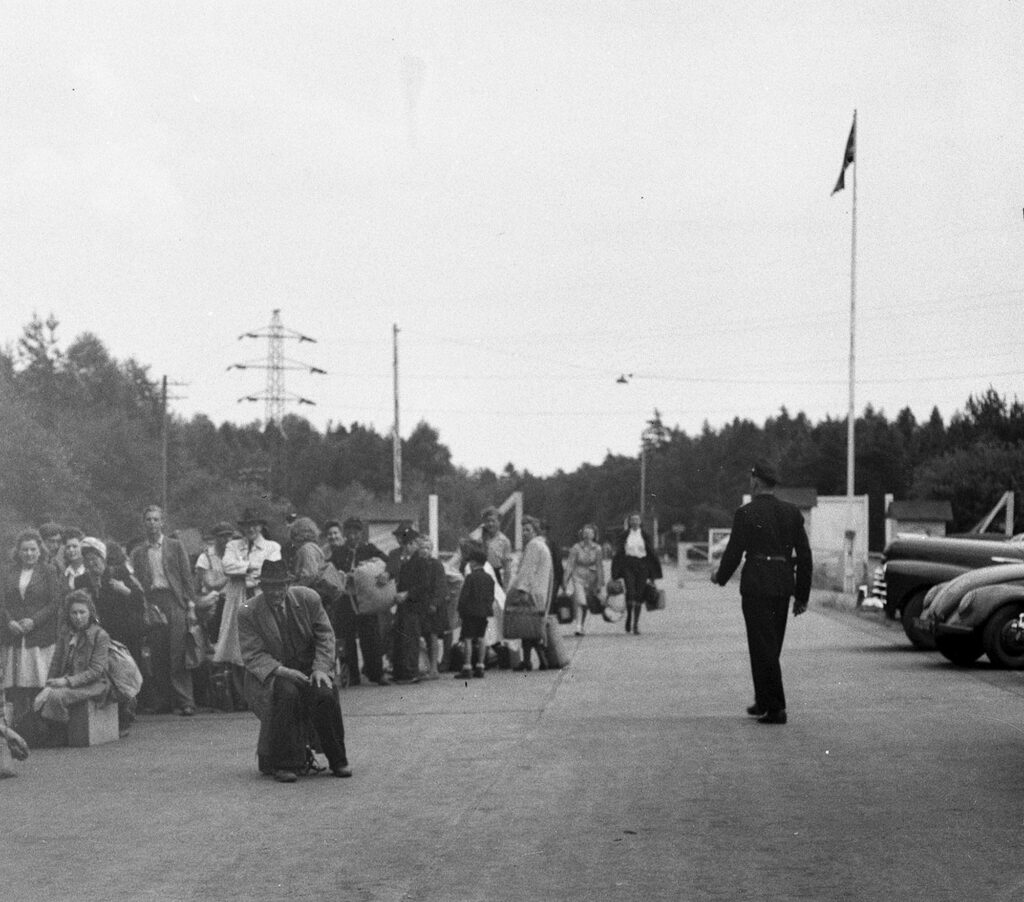 People wait in line at the zone border in Helmstedt. At right, a flagpole with the British flag and a soldier seen from behind.