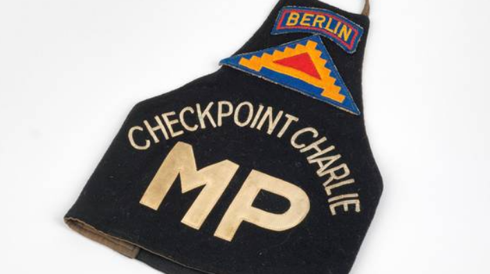 Brassard noir en tissu sur lequel est brodé « Checkpoint Charlie » et les lettres blanches MP, pour Military Police, sont cousues.