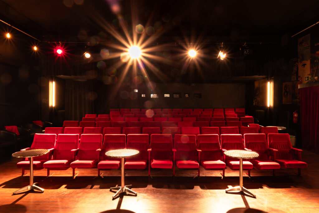 Innenraum eines kleinen Theaters mit roten Sitzplätzen in Reihen, beleuchtet von warmen Scheinwerfern, die von der Bühne in den Zuschauerraum strahlen
