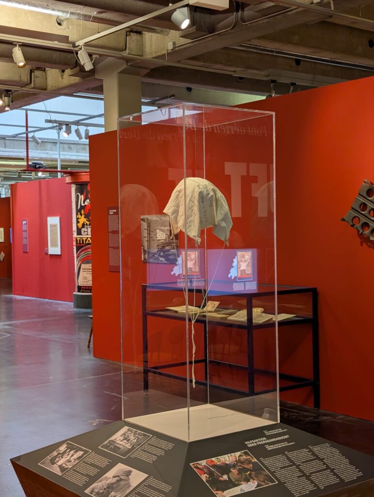 Ausstellungsraum im AlliiertenMuseum, Vitrine mit einem historischen Fallschirm aus der Berliner Luftbrücke in der Ausstellung des AlliiertenMuseums. Im Hintergrund sind rote Ausstellungswände, historische Fotografien und weitere Exponate zu sehen.